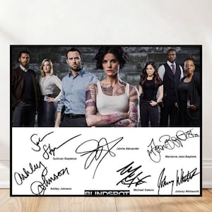 Puede incluir: Un póster enmarcado con una foto del elenco de la serie de televisión Blindspot. La imagen muestra a un grupo de actores, con el título del programa y varias firmas en la parte inferior. Los actores visten diferentes atuendos y el fondo es una pared de ladrillos.