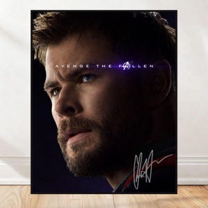 May include: A framed poster featuring a portrait of a man with a beard and blue eyes. The text "AVENGE THE FALLEN" is above the man's head, with the Avengers logo. The poster includes a signature in white.
