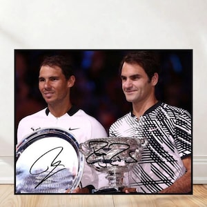 May include: A framed photograph featuring two tennis players holding trophies. One player wears a white shirt, the other a black and white patterned shirt. Both trophies have signatures on them. The photograph is displayed on a wooden surface.