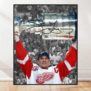 Peut inclure: Photographie encadrée d'un joueur de hockey en maillot rouge et blanc tenant la Coupe Stanley au-dessus de sa tête. Le joueur sourit, avec une foule en noir et blanc en arrière-plan. La photographie est signée.