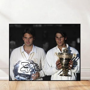 May include: A framed photograph featuring two tennis players holding trophies. One player holds a silver platter, the other a gold trophy. Both are wearing white outfits. The photograph is signed by the players.