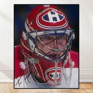 May include: Framed print of a hockey goalie's mask. The red helmet has white and blue details, with the Montreal Canadiens logo. The goalie's face is visible behind the cage. The image has a signature at the bottom.
