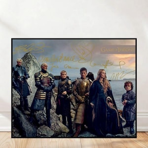 Peut inclure: Une affiche encadrée représentant un groupe de personnes en costumes médiévaux, probablement d'une série fantastique. L'affiche comporte des signatures dorées et les mots "Game of Thrones" en lettres dorées. Le fond montre un paysage côtier.
