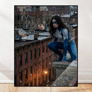 May include: A framed print featuring a person in a superhero costume, crouching on a rooftop. The costume is blue and gray, with a mask. The background shows a cityscape with brick buildings and a cloudy sky. The print has a black frame.