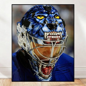 Puede incluir: Primer plano de una máscara de portero de hockey con un diseño de lobo feroz en tonos azules y blancos. La máscara presenta ojos amarillos, dientes afilados y la palabra "TECH". El portero lleva una camiseta azul.