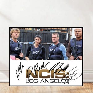 Puede incluir: Un póster enmarcado con el elenco de NCIS: Los Ángeles. La imagen muestra a cuatro personas con chalecos tácticos, con el logotipo del programa y firmas debajo. El fondo incluye una puerta de metal y una pared.