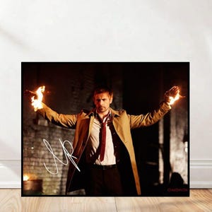 Op de afbeelding: Een ingelijste print met een man in een beige trenchcoat, wit overhemd en rode stropdas, met vlammen in beide handen. De print heeft een handtekening en het woord "Constantine".