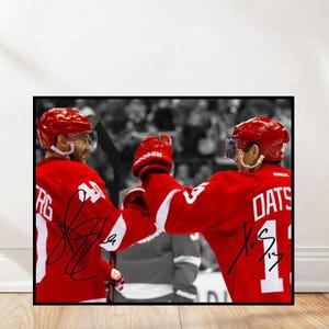 Könnte beinhalten: Gerahmtes Foto von zwei Eishockeyspielern in roten Trikots, die ein Tor feiern. Die Spieler tragen Helme und Handschuhe, das Logo des Teams ist sichtbar. Der Hintergrund ist ein Schwarz-Weiß-Bild der Menge.