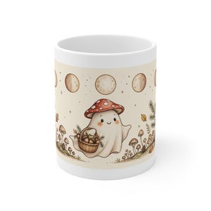 May include: A white ceramic mug featuring a graphic of a small ghost wearing a red and white spotted mushroom cap. The ghost holds a brown woven basket filled with acorns. The design includes moon phases and small forest mushrooms.