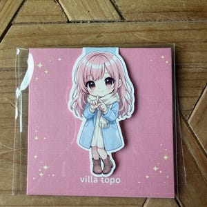 Può includere: Segnalibro rosa con un'illustrazione in stile anime di una ragazza con i capelli rosa, che indossa un cappotto blu e una sciarpa bianca. Il segnalibro è su uno sfondo rosa con il testo "villa topo".