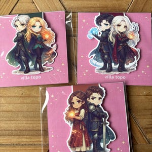 May include: Three stickers with anime-style illustrations of couples in fantasy outfits. Each sticker is on a pink background with the text "villa topo". The characters have different hair colours and hold glowing orbs.