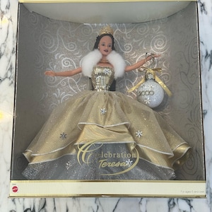 May include: A Celebration Teresa doll in a display box. The doll wears a gold gown with snowflake accents, a white fur stole, and a gold crown. She holds a silver ornament with "2000" on it. The box has the text "Celebration Teresa".
