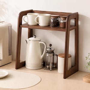 Könnte beinhalten: Ein zweistöckiges hölzernes Arbeitsplattenregal mit dunkelbraunem Finish. Das obere Regal enthält zwei weiße Keramiktassen und zwei Glasgefäße mit Holzdeckeln. Das untere Regal enthält einen weißen Wasserkocher, eine Glas-Pressstempelkanne und einen weißen Behälter.