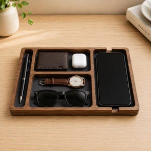 Peut inclure: Un plateau vide-poche rectangulaire en bois avec une finition brun foncé et une doublure en feutre noir, doté de plusieurs compartiments. Il contient un stylo noir, un portefeuille en cuir marron, des écouteurs sans fil blancs, une montre en cuir marron, des lunettes de soleil noires et un smartphone.