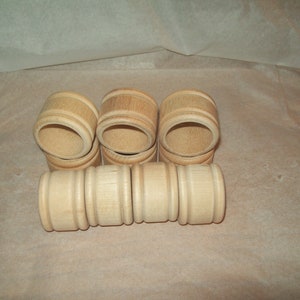 May include: A collection of six wooden napkin rings. The rings are cylindrical with a light wood finish. The design includes horizontal grooves and a smooth, rounded edge. They are arranged on a light brown surface.