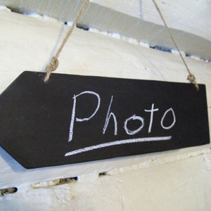 Puede incluir: Un letrero de flecha de pizarra negra con la palabra "Photo" escrita con tiza blanca. El letrero está colgado de una cuerda.