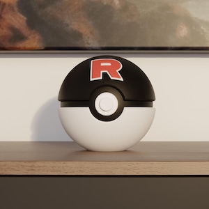 Puede incluir: Una Poké Ball en blanco y negro con el logo "R" rojo en la parte superior. El contenedor esférico está dividido en dos mitades, con una base blanca y una parte superior negra. Un botón blanco está en el centro.