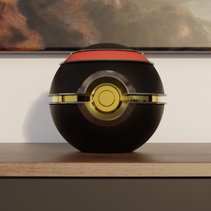 Op de afbeelding: Een zwart en goudkleurig bolvormig object met een rode band. De bol heeft gouden accenten en een cirkelvormig gouden midden. Het staat op een houten oppervlak, met een schilderij op de achtergrond.