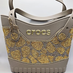 May include: A taupe Crocs tote bag featuring a gold-toned giraffe charm and a giraffe-inspired pattern created with numerous small, sparkling gold and silver rhinestones. The word crocs is embossed in gold lettering on the front centre.