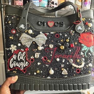 May include: A black Crocs tote bag embellished with numerous black beads, a silver rhinestone bow, a red rose patch, and various charms. The bag features the text "a lil bougie" in white script and the word "crocs" with a red heart.