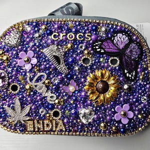 May include: A grey crossbody bag featuring a dense surface of purple and blue beads, gold accents, and various charms including a purple butterfly, a sunflower, a silver leaf, a heart, and the text "crocs" and "ENDIA" in gold lettering.