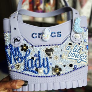 May include: A light purple rubber tote bag featuring the text crocs, Ms Lady, and a lil bougie. The bag is decorated with blue and white rhinestones, pearls, black flower charms, a heart-shaped mirror, and a miniature light blue clog charm.