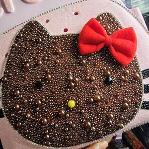 May include: A light pink bag featuring a cat-shaped applique covered in brown beads and pearls. The design includes a bright red fabric bow, two black bead eyes, and a yellow bead nose. The entire cat shape is outlined with a silver rhinestone trim.