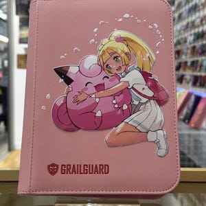 May include: A pink textured binder featuring a graphic of a blonde character hugging a pink Clefairy creature. The cover includes the text GRAILGUARD with a shield logo and scattered white petal illustrations across the front surface.