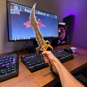 Puede incluir: Una daga decorativa con una hoja curva. La hoja presenta un diseño negro, naranja y dorado. El mango es marrón y negro. La daga se sostiene frente a una configuración de computadora.