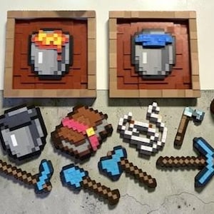 May include: Pixelated art pieces and accessories. Two framed square artworks depict buckets with lava and water. Additional items include a bucket, backpack, spider web, pickaxes, axes, and a torch.