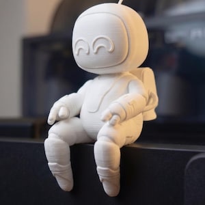 May include: A white 3D printed figurine of a small astronaut with a rounded helmet, curved eyes, and a backpack. The figure is depicted in a seated position with legs dangling, featuring visible layer lines from the additive manufacturing process.