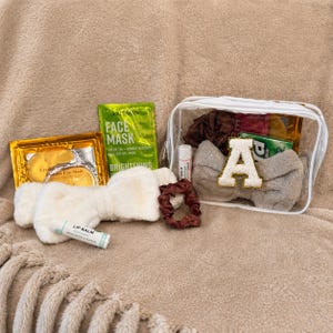 May include: A clear cosmetic bag filled with beauty products, including a headband with an "A" patch, lip balm, face mask, and eye mask. A brown scrunchie and a fluffy white headband are also included. The items are arranged on a soft, beige surface.