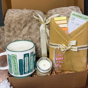 May include: A gift box containing a beige textured throw blanket tied with a ribbon, a white ceramic mug with green book spine illustrations, a small candle in a glass jar, and a wrapped book with a reading list card and a tassel.