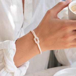 Op de afbeelding: Een verfijnde zilverkleurige armband met een centraal knoopontwerp. De armband bestaat uit twee dunne, parallelle metalen strengen die samenkomen in een klein, gedetailleerd knoopje, wat zorgt voor een elegante uitstraling om de pols.