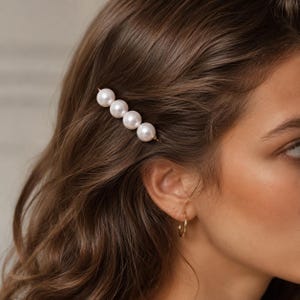 May include: A hair clip featuring four round, white, lustrous pearls arranged in a straight horizontal line on a thin gold-toned metal bar. The accessory is secured in brown hair, positioned above the ear, and paired with a small gold hoop earring.