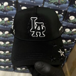 以下が含まれることがあります： 黒いベースボールキャップ。白い刺繍で「LA」の文字と星のデザイン。つばはカーブしており、星のアクセントも。耐久性のある生地で作られています。