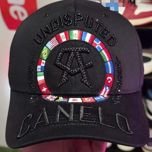 Puede incluir: Gorra de béisbol negra con la palabra "UNDISPUTED" bordada en la parte delantera. La gorra presenta un diseño circular con banderas y el nombre "CANELO" bordado en la visera. La gorra tiene una visera curva.