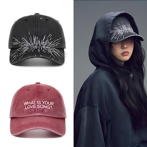 Puede incluir: Una gorra de béisbol negra con un gráfico de estallido blanco y una gorra roja bordada con el texto blanco WHAT IS YOUR LOVE SONG? Ambas gorras tienen una visera curva y una corona estructurada con un botón en la parte superior.