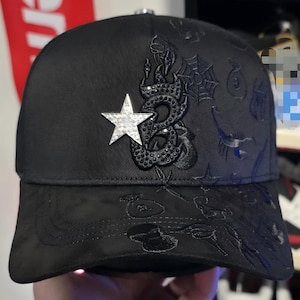 May include: Black baseball cap with a curved brim. The cap features a silver star and a decorative snake design. The cap has embroidered details and a metal clasp on the back.