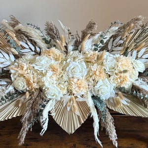 May include: A large floral arrangement featuring cream-colored roses, carnations, and dried pampas grass. The arrangement includes fan-shaped palm leaves and various other dried elements, creating a natural, textured look. The centerpiece is set on a wooden table.