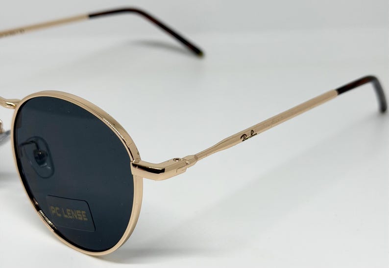 Puede incluir: Gafas de sol de metal dorado con lentes oscuras. Las monturas y varillas redondas son doradas, con un logotipo sutil en la varilla. Las lentes son oscuras y el dise&ntilde;o es cl&aacute;sico y elegante.