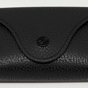 Puede incluir: Estuche de gafas de cuero negro con superficie texturizada. El estuche tiene una parte superior curvada y un cierre a presi&oacute;n. Un logotipo Ray-Ban dorado y negro es visible en la parte delantera.