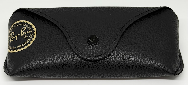 Puede incluir: Estuche de gafas de cuero negro con superficie texturizada. El estuche tiene una parte superior curvada y un cierre a presi&oacute;n. Un logotipo Ray-Ban dorado y negro es visible en la parte delantera.