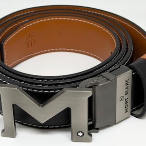 Puede incluir: Cinturón de cuero negro con hebilla plateada con un gran diseño de "M". El cinturón tiene un interior de cuero marrón y costuras blancas. La hebilla está marcada "MONT BLANC". Talla del cinturón: 115 cm.