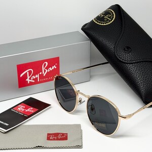 Puede incluir: Gafas de sol Ray-Ban con montura dorada y lentes oscuras, estuche de cuero negro, pa&ntilde;o de limpieza gris y caja plateada con el logotipo de Ray-Ban. Las gafas de sol y los accesorios se muestran sobre una superficie blanca.