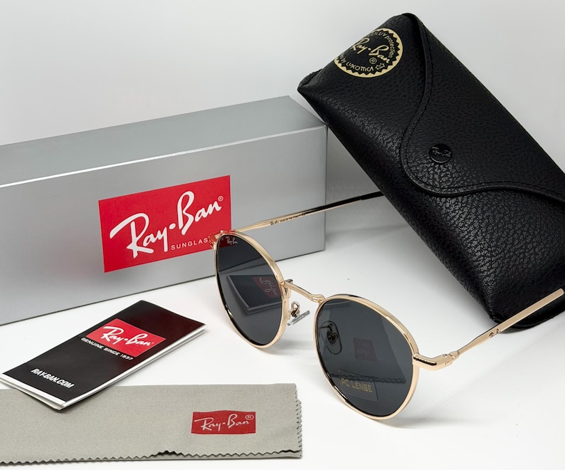 Puede incluir: Gafas de sol Ray-Ban con montura dorada y lentes oscuras, estuche de cuero negro, pa&ntilde;o de limpieza gris y caja plateada con el logotipo de Ray-Ban. Las gafas de sol y los accesorios se muestran sobre una superficie blanca.