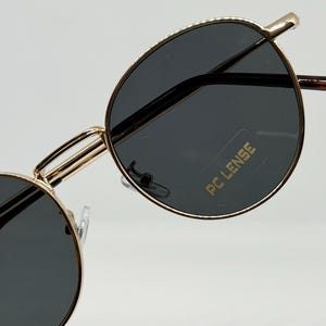 Puede incluir: Primer plano de gafas de sol con montura dorada y lentes grises oscuras. Las gafas tienen una forma redonda cl&aacute;sica y una montura dorada fina. Las lentes tienen el texto "PC LENSE" impreso.