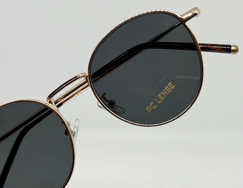 Puede incluir: Primer plano de gafas de sol con montura dorada y lentes grises oscuras. Las gafas tienen una forma redonda cl&aacute;sica y una montura dorada fina. Las lentes tienen el texto "PC LENSE" impreso.
