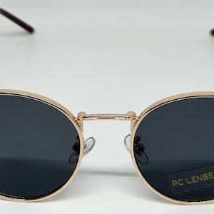 Puede incluir: Gafas de sol con montura dorada y lentes oscuras. Las lentes ovaladas est&aacute;n engastadas en una montura de metal dorado con puente y patillas. El logotipo de Ray-Ban es visible en la lente izquierda. Las gafas tienen una pegatina PC LENSE.