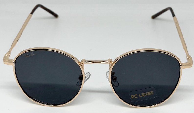 Puede incluir: Gafas de sol con montura dorada y lentes oscuras. Las lentes ovaladas est&aacute;n engastadas en una montura de metal dorado con puente y patillas. El logotipo de Ray-Ban es visible en la lente izquierda. Las gafas tienen una pegatina PC LENSE.